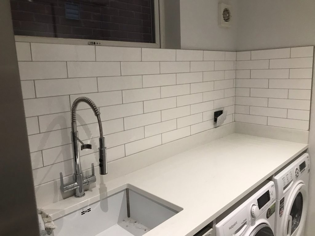 Utility room walls tiled in a decorative white 300mm long tile showing sink splash and worktop.